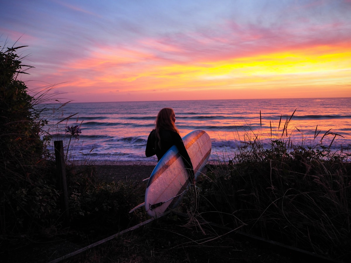 Woman holding surfboard at sunset beach - best swimwear for water sports 2026