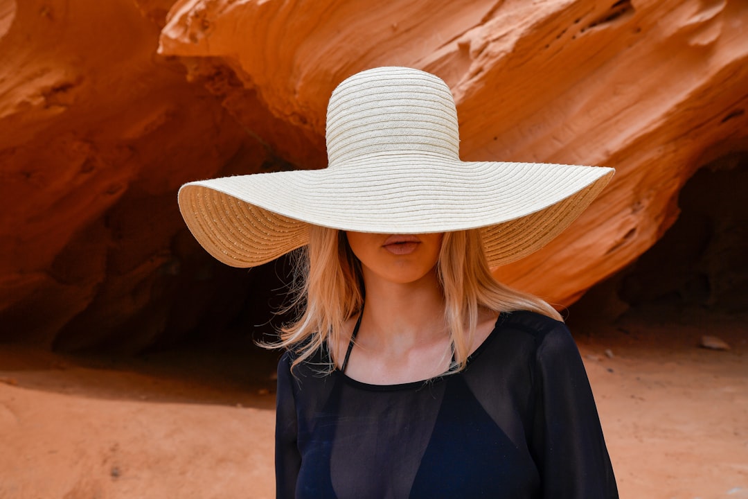 A young blonde woman wearing a light straw hat stands at the entrance of a red sandstone cave in Mallorca, Spain.