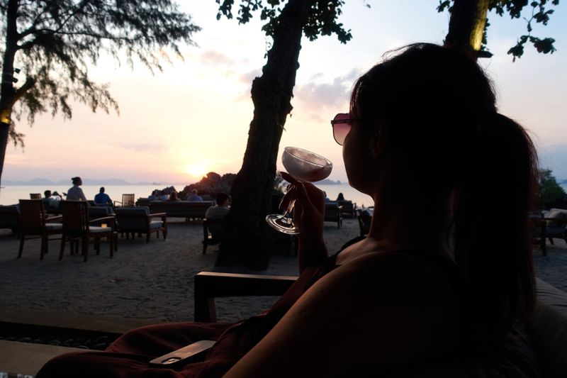 Woman in resort wear enjoying cocktail at beach bar during sunset, beach to bar outfit transition style