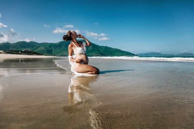 Pregnant woman in white maternity bikini on a tropical beach with mountain backdrop