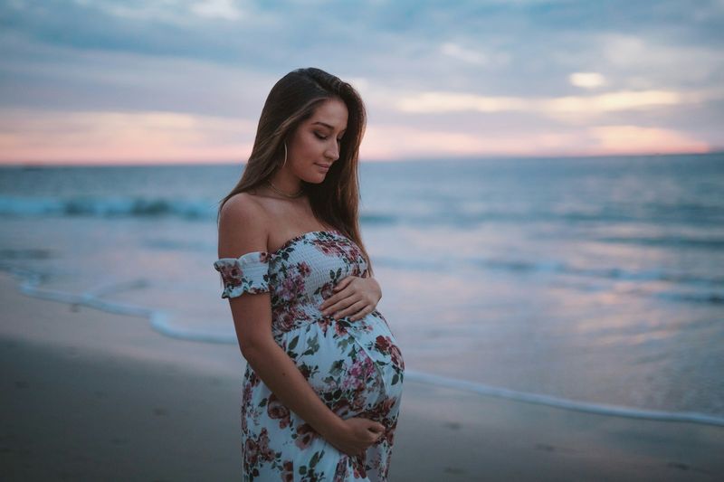 Pregnant woman in a floral maternity cover-up dress worn over a bikini on the beach at sunset