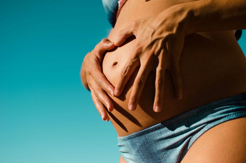 Close-up of a pregnant belly with hands forming a heart over a high-waisted maternity bikini bottom