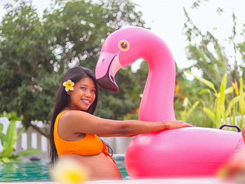 Pregnant woman in an orange maternity swimsuit floating with a pink flamingo pool float