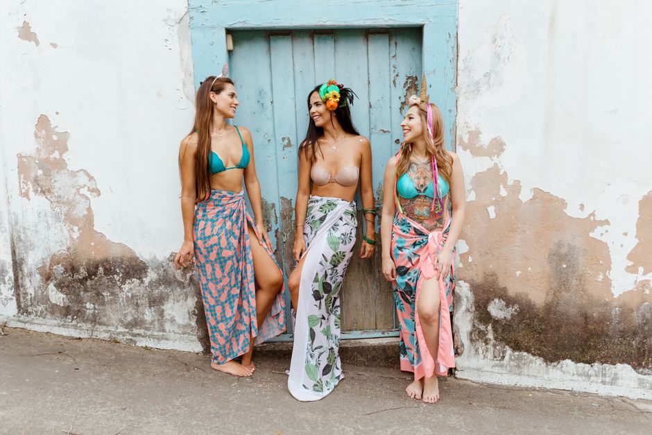 Three women in summer attire smiling by a rustic wall, showcasing vibrant sarongs and floral headpieces.