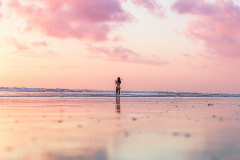 Woman in a three-piece swimsuit standing at the shoreline at sunset
