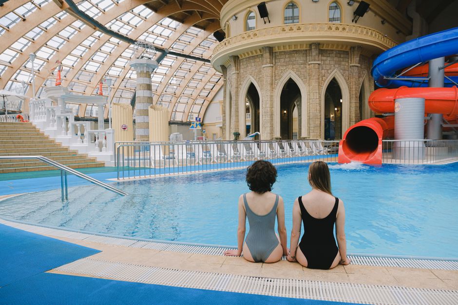 Two women in swimsuits sitting by an indoor pool with water slides.