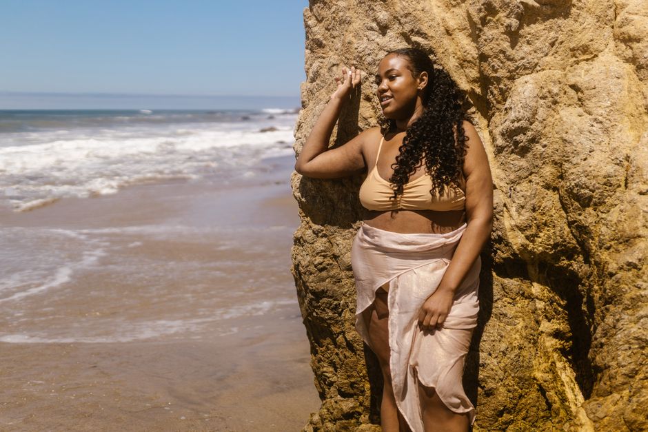 Young woman with curly hair leaning against a rock by the ocean, enjoying a sunny day at the beach.