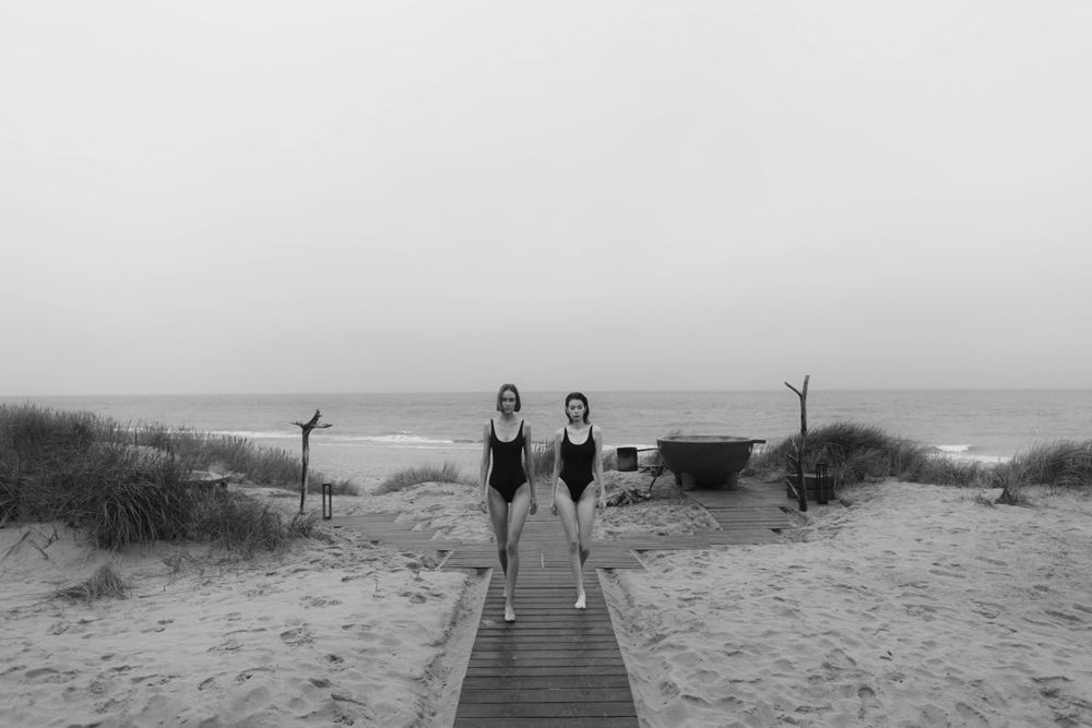 two women in one-piece swimsuits on the beach comparing swimsuit styles