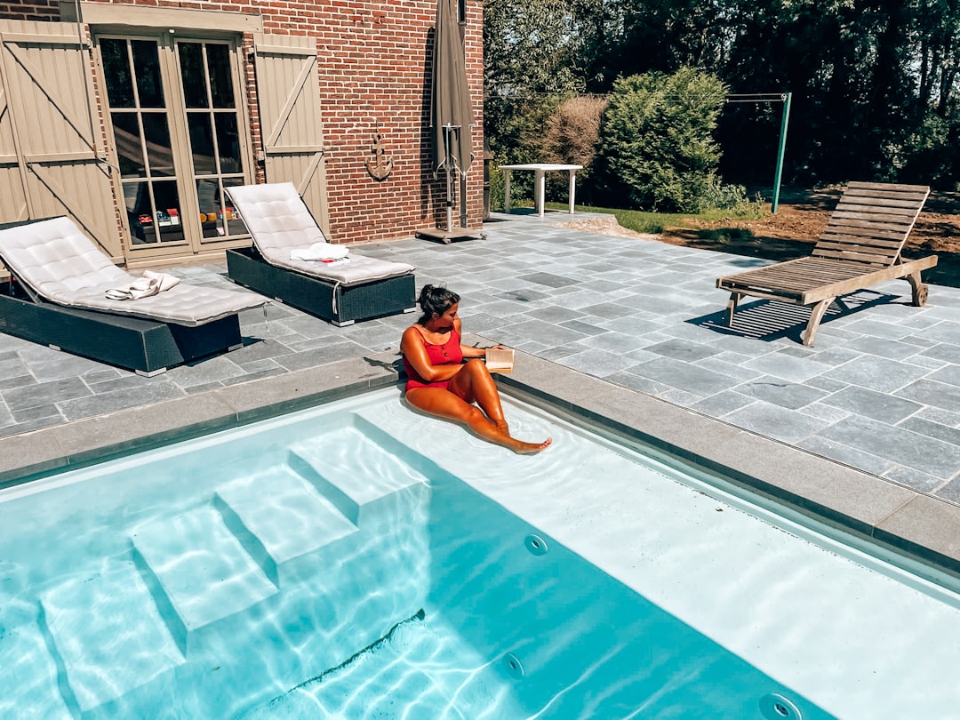A woman in a red swimsuit sits by a modern backyard pool, reading in the sun. The scene evokes a peaceful summer moment of se