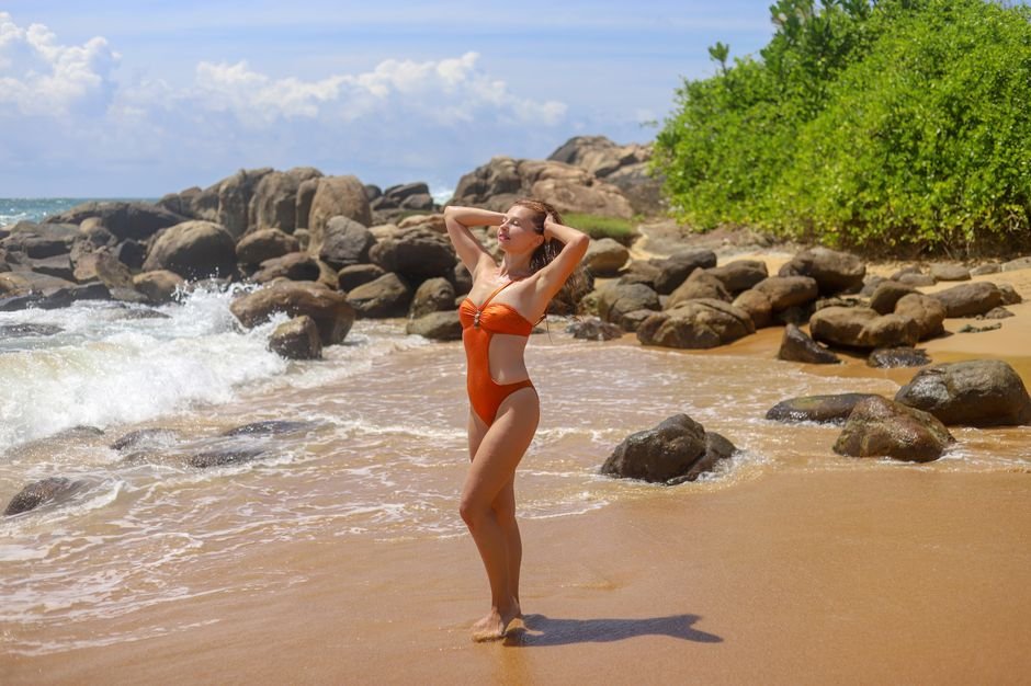 A woman enjoys a sunny day on a rocky beach, wearing an orange swimsuit.