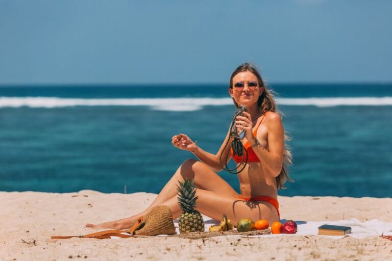 Woman in bikini relaxing on a sunny beach with tropical fruits and a refreshing drink.