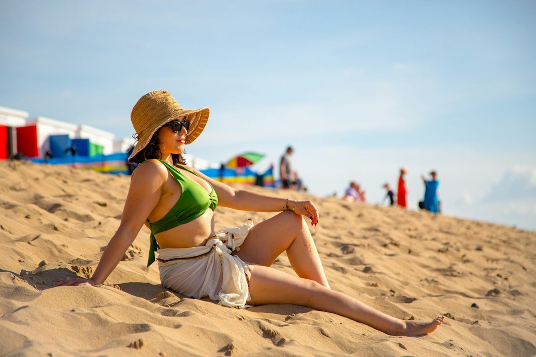 A woman in a green bikini and straw hat sitting on a sandy beach