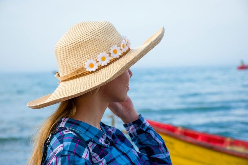 Woman in a wide-brim sun hat standing near the water with a boat in the background